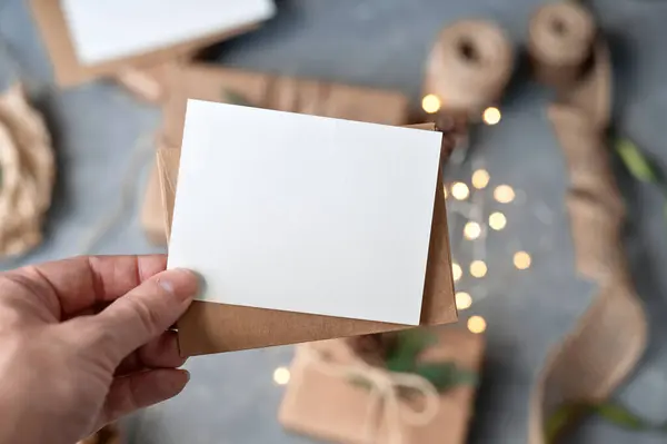 Elinizde boş bir kağıt maket modeli ve bulanık masa arkaplanının üzerinde Noel hediyesi kutuları ve parlak çelenk ışıkları olan bir zarf var. Noel tebrik kartı veya davet şablonu.