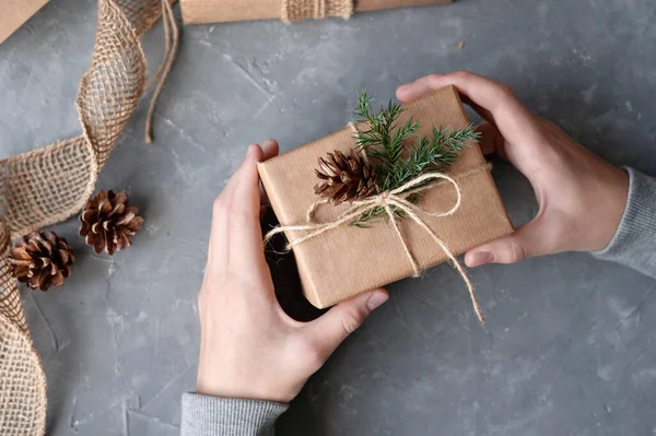 Person\'s hands holding brown crafted gift box decorated with pine branch and cone over gray table background. Sustainable aesthetic Christmas celebration, presents preparing concept.