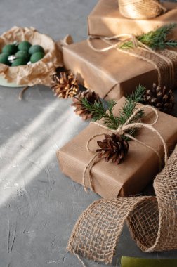 Zarif kırsal Noel kutlaması, el yapımı kahverengi kağıt, çam dalı ve koni ile süslenmiş hediye kutuları, gri masa arkasında doğal güneş ışığı ve gölgeler. Yaşam tarzı tatil kompozisyonu.
