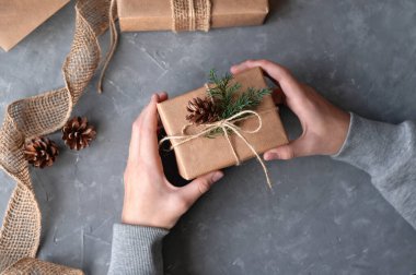 Noel hediyesi kutusunu tutan eller kozalaklı koniler ve dallarla süslenmiş, el yapımı kağıda sarılmış, gri masa arkasında..