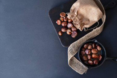 Kavrulmuş kestane kestaneleri siyah ahşap tahtada ve tavada, fotokopi aletiyle birlikte koyu arka planda. Geleneksel kış yemekleriyle estetik düzlük.