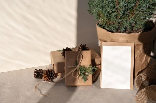 Crafted gift boxes, empty paper card mockup, juniper in pot on table with beige linen tablecloth, with natural sunlight shadows, lifestyle holiday winter composition, sustainable Christmas decoration.
