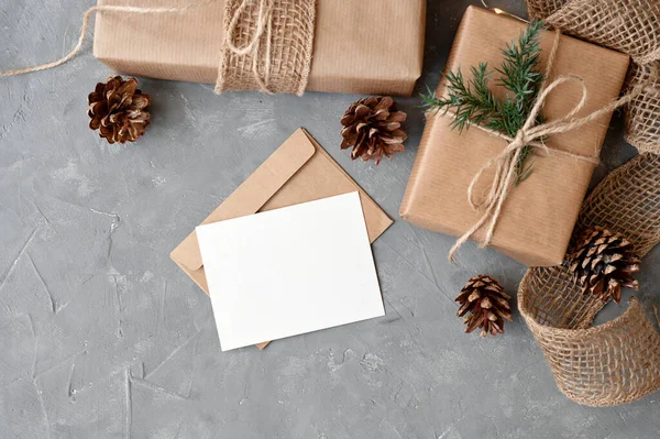 Christmas presents and postcard on table, aesthetic sustainable holiday celebration template. Blank paper card mockup, brown crafted gift boxes on gray table background, pine cones, twigs decoration.