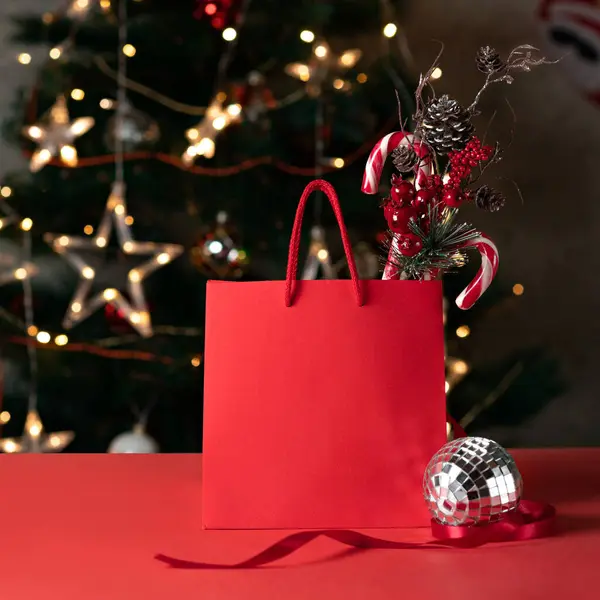 赤い輝くショッピングバッグモックアップ装飾的な松の木の枝 キャンディー缶 ぼやけたクリスマスツリーガーランドライトと赤いテーブルのミラーボール 休日の買い物販売のためのビジネスブランドのテンプレート — ストック写真