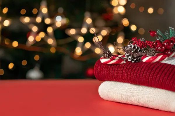 Boş fotokopi alanı olan kırmızı masa üstü, kırmızı ve beyaz örgü süveterler, çam ağacı dalı süslemesi, şeker kamışı, arka planda bulanık Noel ağacı ışıkları. Mağaza ürün yerleştirme için tatil şablonu.