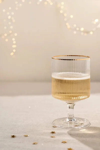 Wineglass with sparkling wine alcohol drink on beige linen tablecloth with natural shadows and gold confetti, blurred garland lights on background. Aesthetic New Year, wedding, birthday background.
