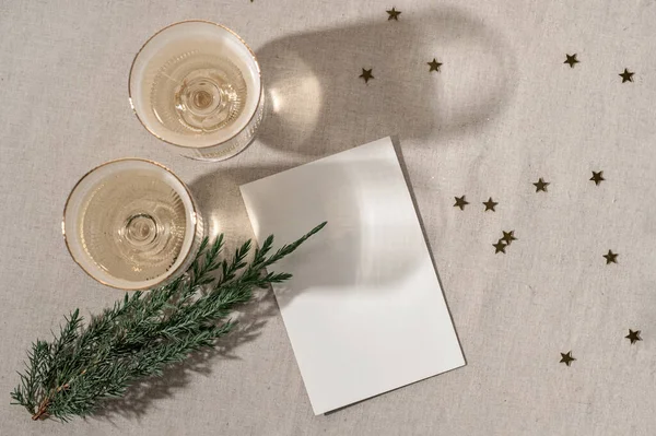 Minimal aesthetic holiday template, blank paper card mockup, wineglasses with sparkling wine, gold confetti and juniper branch on beige linen background, top view, flat lay.