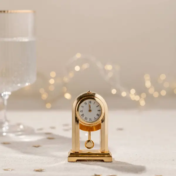 New Year holiday party celebration time. Vintage gold clock showing twelve o\'clock, midnight, on beige linen tablecloth, blurred wineglass with sparkling wine, garland lights on background.