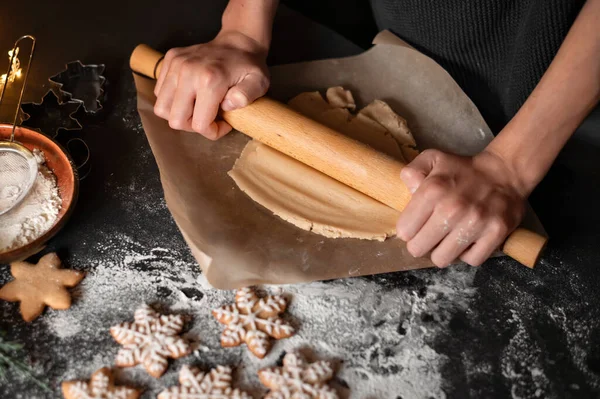 Hands holding rolling pin, rolling dough on parchment on black table background with flour, preparing for gingerbread cookie baking. Traditional Christmas home bakery. Soft selective focus, lifestyle.