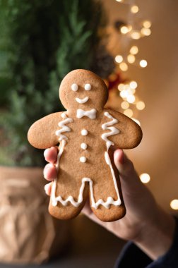 Elinde zencefilli kurabiye, arka planda bulanık Noel ağacı ve çelenk ışıkları. Aile kış bayramı kutlaması, geleneksel çocuk Noel ziyafeti. Yumuşak seçici odak.