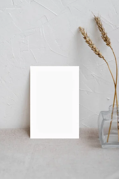 Minimalist home interior poster or picture frame design template, blank paper card mockup on neutral beige table, vase with dried meadow grass, empty white plaster textured wall background.