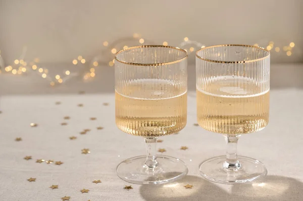 Two wineglasses with sparkling wine on beige linen tablecloth with confetti and light shadows, blurred garland lights on background. New Year, anniversary, wedding or birthday celebration