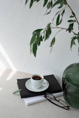Beyaz kahve fincanı, kitaplar ve gözlükler, bej masa üzerinde bitki bulunan yeşil vazo, güneş ışığı lekeli beyaz alçı duvar arkaplanı. Estetik kış, ticari markalaşma için hala hayat.