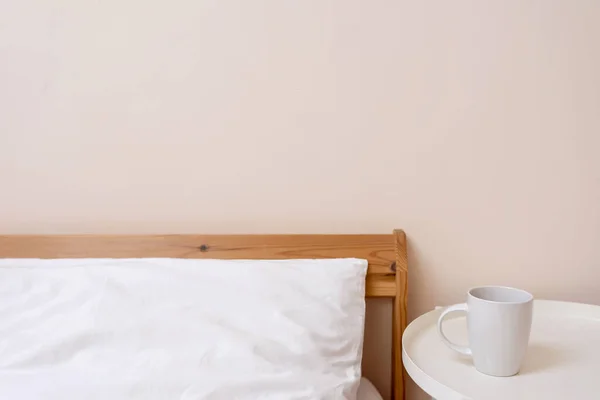 Lifestyle minimal bedroom interior detail, bed with white pillow. coffee cup on bedside table, empty peachy neutral wall background.