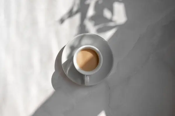 Minimal aesthetic lifestyle coffee concept, saucer and cup with milky coffee on marble gray table background with natural geometric sunlight shadow, good morning business branding backdrop.