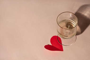 Minimum Sevgililer Günü kutlaması konsepti, köpüklü şarap kadehi ve nötr şeftali rengi arka planda sert gölgeler altında kırmızı kağıt kalp şeklinde sevgililer günü kartı..