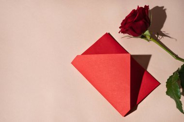 Lüks kırmızı aşk mektubu, randevu davetiyesi, tebrik kartı ya da hediye. Kırmızı kağıt zarf ve gül çiçeği pastel renksiz bej arka planda, düz yerde..