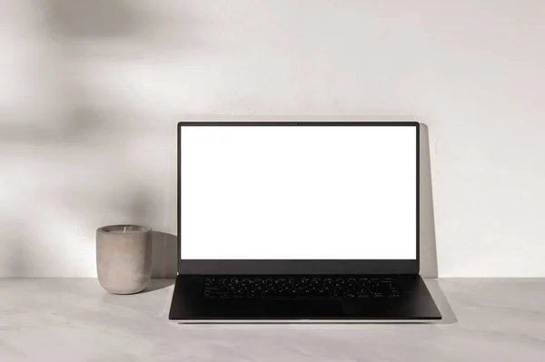 Laptop computer with white empty screen mockup, candle on neutral beige marble table, warm white textured plaster wall background with soft sunlight shadows, minimal business design template.
