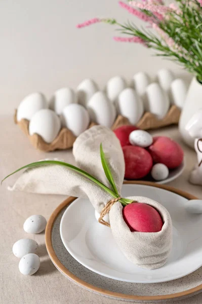 Aesthetic Easter holiday decor for table setting, pink colored egg decorated with bunny ears napkin on white plate, blurred eggs in box on background, spring flowers in vase, soft natural sun light.
