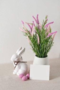 Fotokopi alanı, beyaz porselen tavşan heykelciği, vazoda pembe bahar çiçekleri ve doğal bej keten arka planda yumurtalar, Paskalya bayramı ev dekorasyonu, tebrik kartı şablonu..
