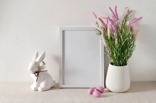 Blank white picture frame mockup, flowers in vase, rabbit figurine, pink candy eggs on neutral beige table background, soft natural sun light shadows, Easter home decor, holiday poster template.