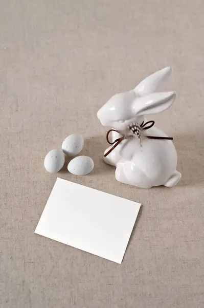 Minimalist aesthetic Easter still life, blank paper card mockup, rabbit figurine, small eggs on neutral beige linen cloth background.