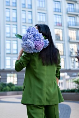 Moda yeşili ceket ve pantolon giymiş uzun saçlı yetişkin esmer kadın şehir binalarında büyük bir buket ortanca çiçeği tutuyor, arka plan, seçici odaklanma, yaşam tarzı.