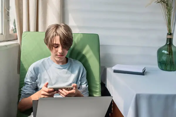 Youth and teenagers gadget and internet addiction concept, handsome teenager boy with laptop indoor in bright sunlight, holding smartphone in hands and looking on screen, lifestyle.