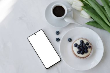 Boş ekran maketli cep telefonu, kahve fincanı, panna cotta tatlı ve beyaz masa arkaplanındaki çiçekler doğal güneş ışığı, estetik ticari markalaşma şablonu, küçük işletme konsepti.