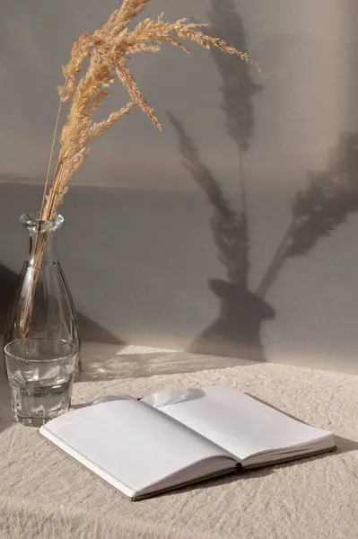 .Open book and vase with dried grass on table with beige linen tablecloth, summer boho still life, aesthetic sustainable home interior in earthy colors.