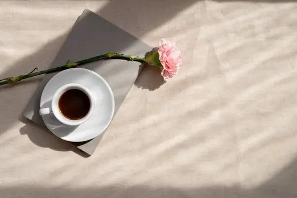 Minimalist aesthetic spring or summer still life, coffee cup, notebook and carnation flower on beige linen table background with abstract lifestyle sunlight shadows.