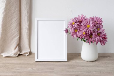 Boş beyaz fotoğraf çerçevesi veya poster modeli, açık bej ahşap masa üzerindeki pembe çiçekli vazo, keten tekstil perde ve beyaz duvar arkaplanı, İskandinav tarzı estetik natürmort..
