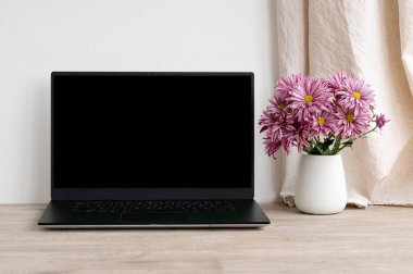 Siyah ekranlı dizüstü bilgisayar, nötr bej ahşap masadaki çiçekli vazo, beyaz beton duvar arka planı, estetik konforlu ofis çalışma alanı.