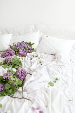 Dağınık beyaz yatak odasındaki leylak çiçekleri, çiçek açan en az İskandinav beyazı yatak odaları, pamuk çarşafların üzerindeki yapraklar, battaniye ve yastıklar, çiçeklerin markalaştırma şablonu.