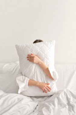 İyi uyku, rahat yatak konsepti, yastığı kucaklayan insan, yastığın arkasına saklanan, yatakta oturan, asgari estetik beyaz, tarafsız yatak odası, İskandinav tarzı..