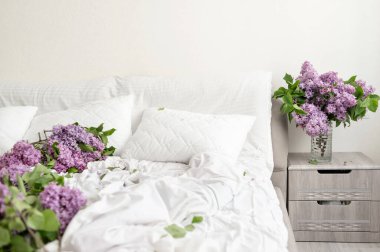 Estetik romantik yatak odası iç kısmı, beyaz buruşmuş çarşaf döşeli yapılmamış yatak, komodindeki vazoda leylak çiçekleri, dağınık leylak çiçekleri, çarşaflarda yapraklar ve yapraklar..
