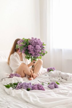 Beyaz havadar yatak odasında, yumuşak doğal ışık, leylak çiçeği buketi, arkasında saklanan yüz, yatakta dağınık çiçek yaprağı, estetik çiçekli ergenlik konsepti..