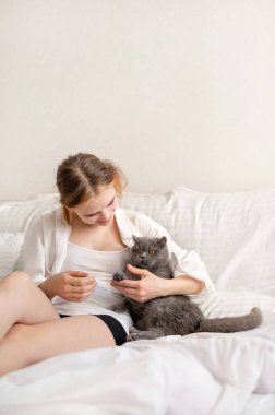 Teenager girl taking care about her gray cat, sitting on bed together, embracing and stroking pet. clipart