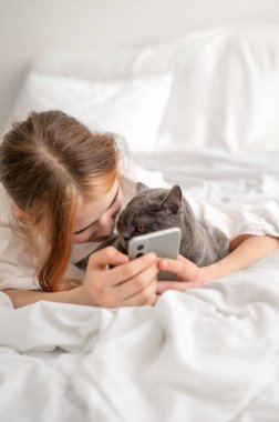 Genç kız, cep telefonu ve yaşam tarzı gibi sosyal medya araçları kullanarak kedisiyle birlikte yatakta oyun oynuyor..