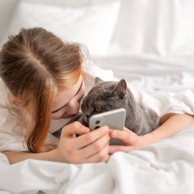 Kız cep telefonu kullanırken kediyle oynuyor, yatakta yan yana yatıyor, beyaz yatak odası..