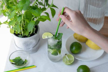 Ev yapımı tatlı yaz mojito kokteyli, elinde bardakta saman tutan kişi, yeşil limon, beyaz masa arkasında nane ve yeşil limon, üst manzara..