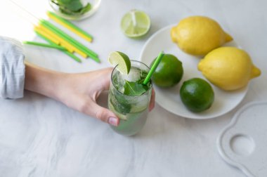 Buzlu mojito kokteyli, yeşil limon, sarı limon, beyaz mermer masa arka planında pipet içen kişi, üst görüş, doğal güneş ışığı, yumuşak seçici odak.