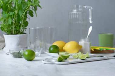 Tam ve dilimlenmiş yeşil limon, limon, nane, boş bardaklar, şeker ve karbonatlı su beyaz mermer masa zemininde, ev yapımı mojito kokteyli yapmak için malzemeler.