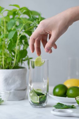 Ev yapımı mojito kokteyli hazırlama süreci, dilimlenmiş yeşil limonları nane yapraklı bardağa koyma, masada mojito malzemeleri, tarafsız gri arka plan, yumuşak doğal ışık..