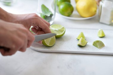 Kesme tahtasında limon dilimleyen eller, limon, beyaz masa arka planında nane, yumuşak doğal ışık, taze sulu narenciye meyvesi, mojito tarifi için malzemeler hazırlamak..