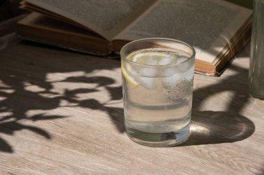 Buzlu ferahlatıcı limonata ya da limonata katılmış su, ahşap masa arkasında açık kitap. Estetik güneş ışığı gölgeleri, sağlıklı yaz içeceği, kopyalama alanı..