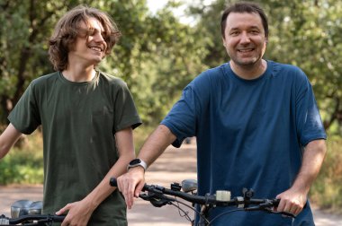 Babası, yaz parkında bisiklet sürerken, oğlunun cep telefonuyla fotoğrafını çekiyor. Aile aktiviteleri ve birliktelik..