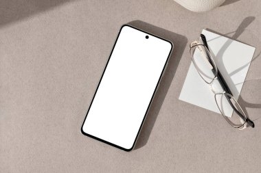 Boş beyaz ekranlı cep telefonu, gözlükler, kahverengi masa arkaplanındaki kağıt kart, estetik güneş ışığı gölgeleri, minimalist ticari marka tasarım şablonu, düz yatış..