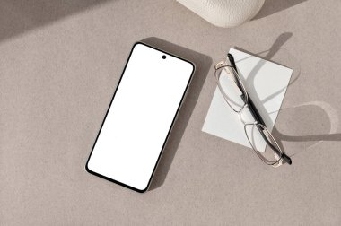 Beyaz ekranlı akıllı telefon, gözlükler, nötr kahverengi masa arkaplanı üzerine kağıt kart doğal güneş ışığı gölgeleri, minimalist ofis çalışma alanı, ticari marka tasarım şablonu.