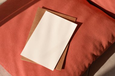 Boş kağıt maketi, terracotta kırmızı desenli yastık zemininde doğal güneş ışığı gölgeleri, rahat ev ayrıntıları, sonbahar kartpostalı ya da davetiye tasarımı şablonu içeren el yapımı bir zarf..